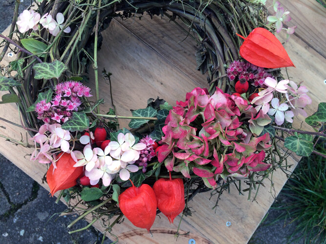 Corona otoñal hecha a mano con flores rosas, farolillos naranjas y hojas verdes sobre una tabla