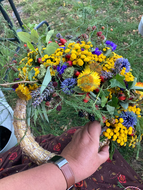 Corona otoñal a medio atar con flores amarillas y azules, gramíneas y bayas sobre base de paja